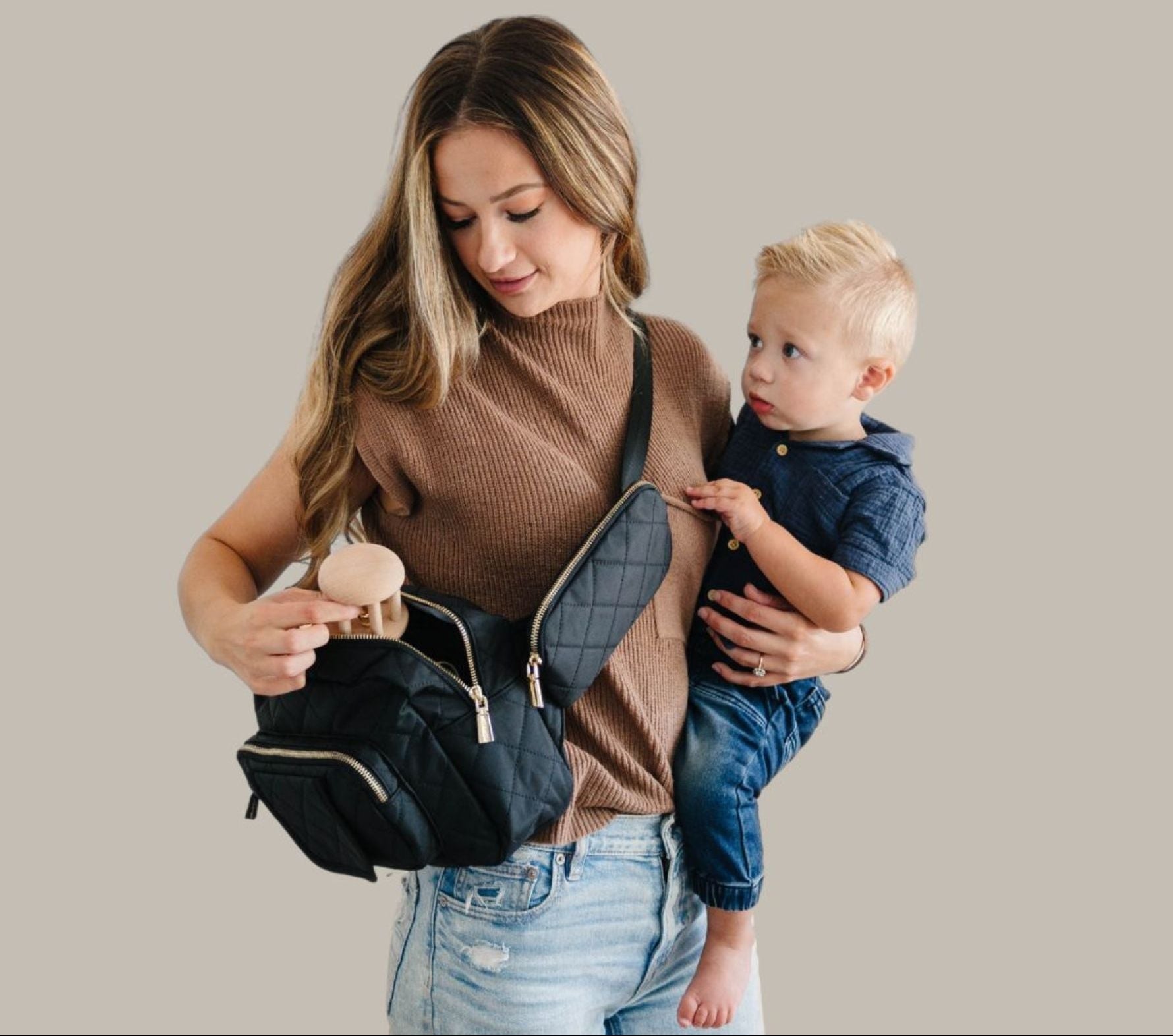 Mom holding a baby and a black min diaper bag against a beige background
